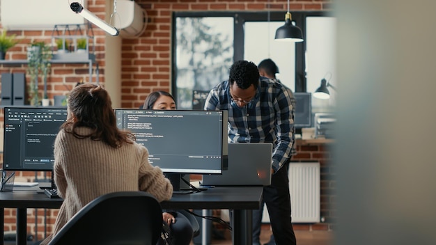 database programer writing code in front of multiple computer screens displaying artificial intelligence algorithm. developer coding database while colleagues doing teamwork in background.