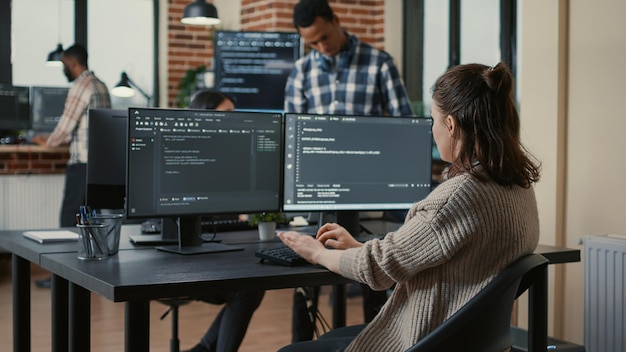 focused software developer writing code looking at multiple computer screens displaying machine learning algorithm. programer coding user interface while colleagues doing teamwork in background.